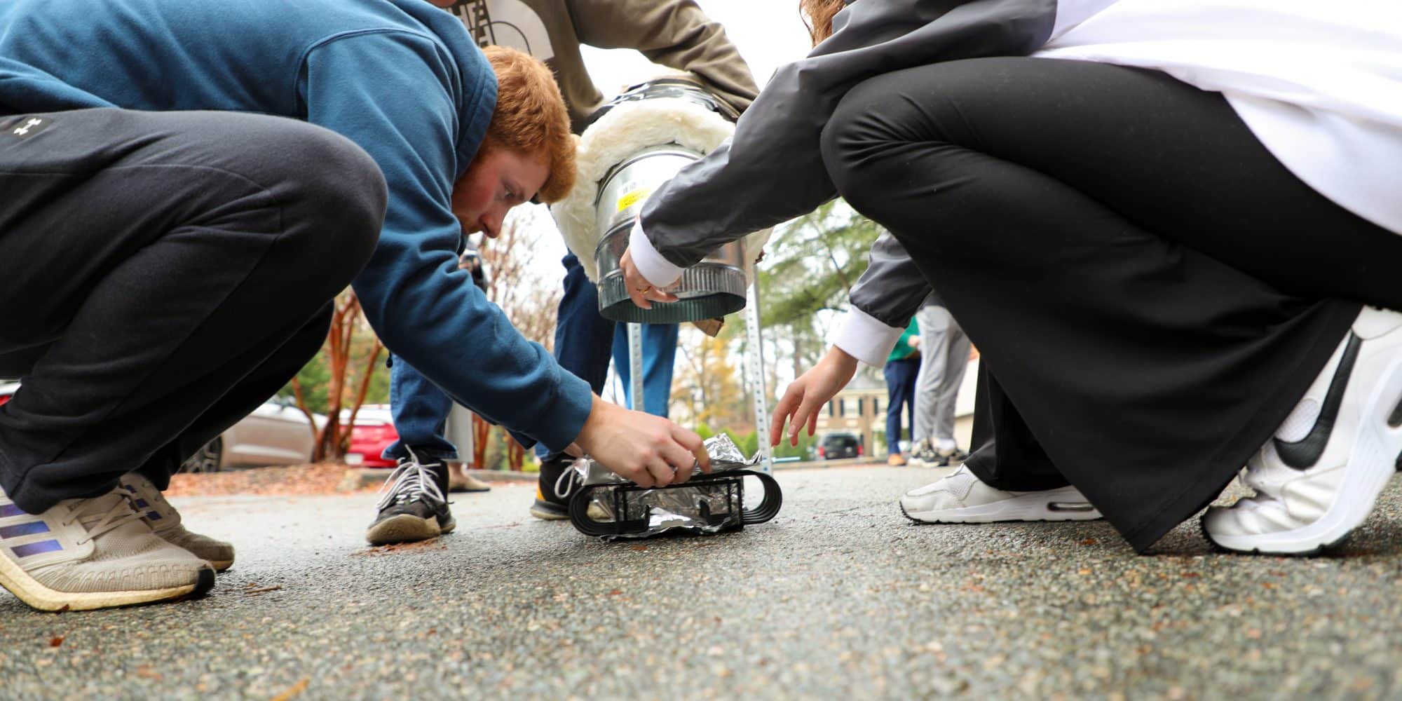 Randolph-Macon engineering students outdoors assembling a small robotic device on a pathway, surrounded by others observing the engineering process.